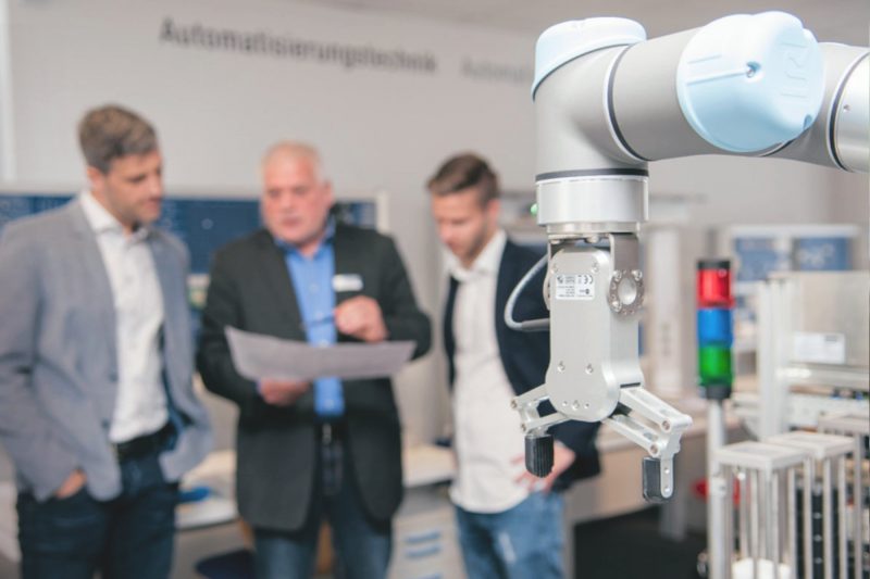 im Vordergrund steht ein Roboterarm, im Hintergrund wird von drei Menschen über diesen arm diskutiert
